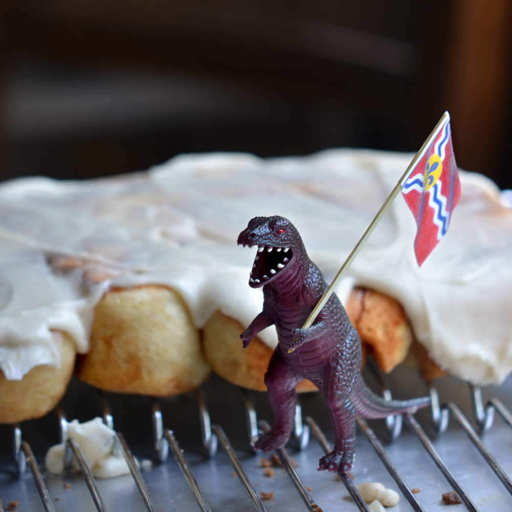 Henry/Hank holding a St. Louis flag standing in front of cinnamon rolls on a baking rack.